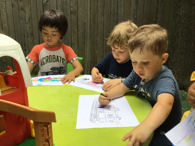 Three young boys sit at a green table coloring line drawings with crayons, with a red and white toy structure visible on the left.