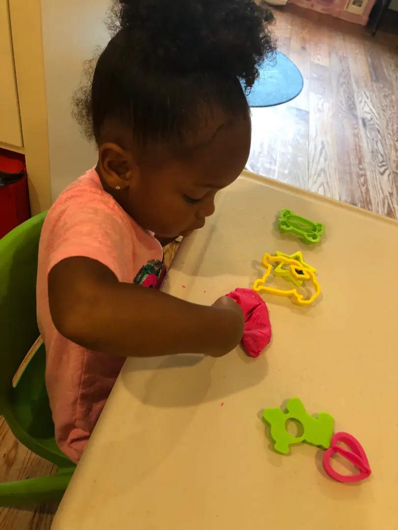 A young child in a pink shirt sits at a table and presses red play dough with a green shape cutter while other plastic cutters rest nearby.