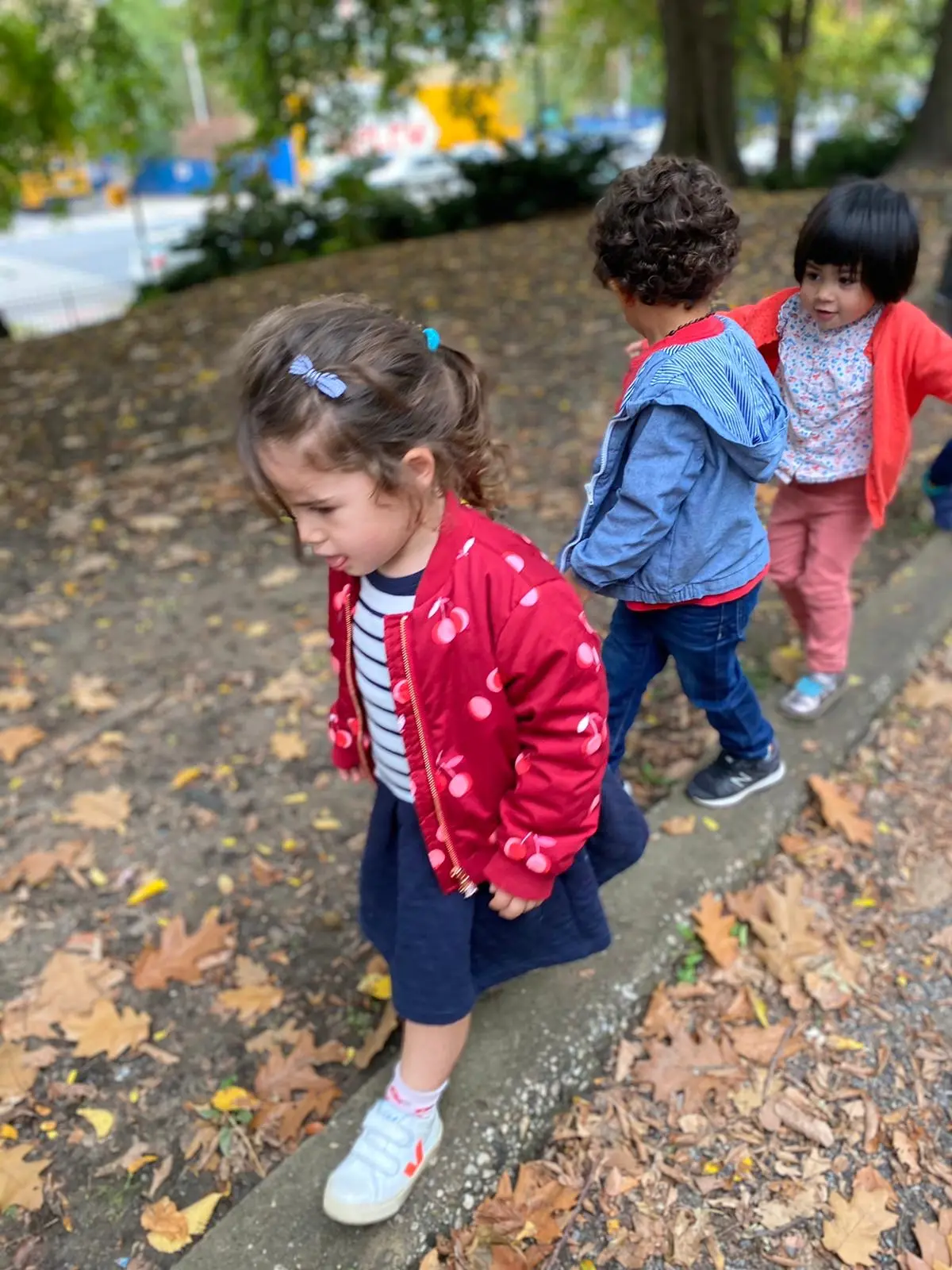Three young children walk along a concrete curb scattered with fallen leaves, with the child in front wearing a red cherry-patterned jacket and looking down.