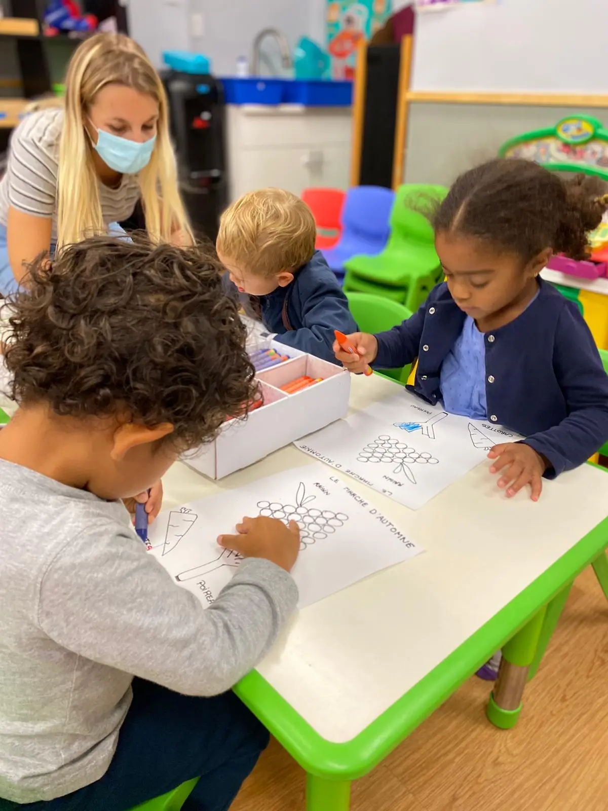 Three children color printed worksheets with crayons at a green table while an adult in a mask watches over them.