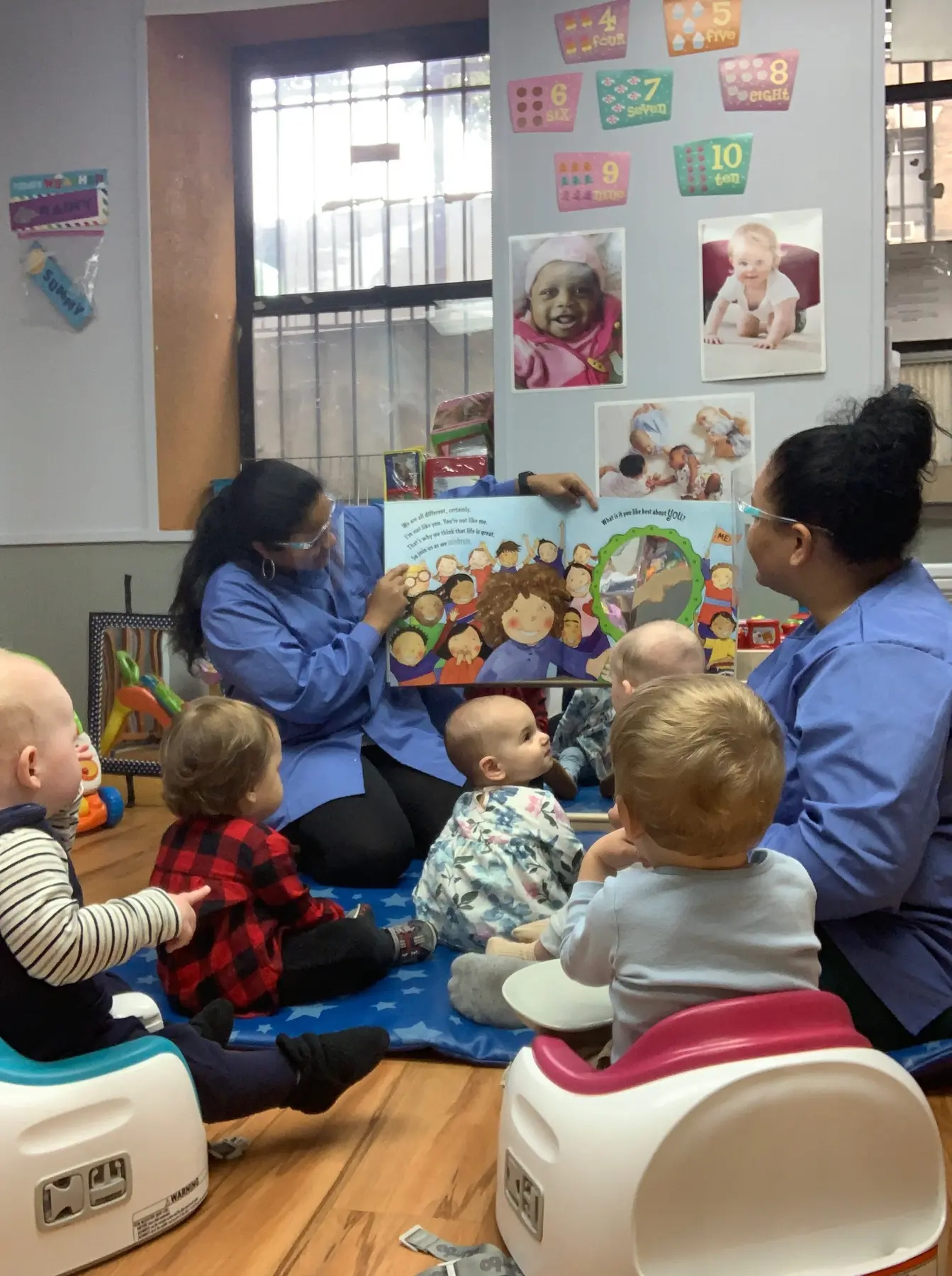 Two adults in blue shirts sit on a blue mat holding a picture book for five seated babies and toddlers in a room with number posters and baby photos on the wall.