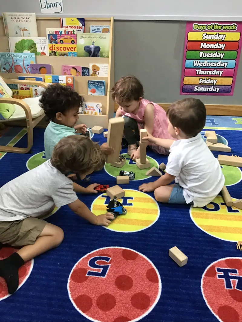 Four children sit on a patterned rug building with wooden blocks and toy cars in front of a bookshelf and a days-of-the-week poster.