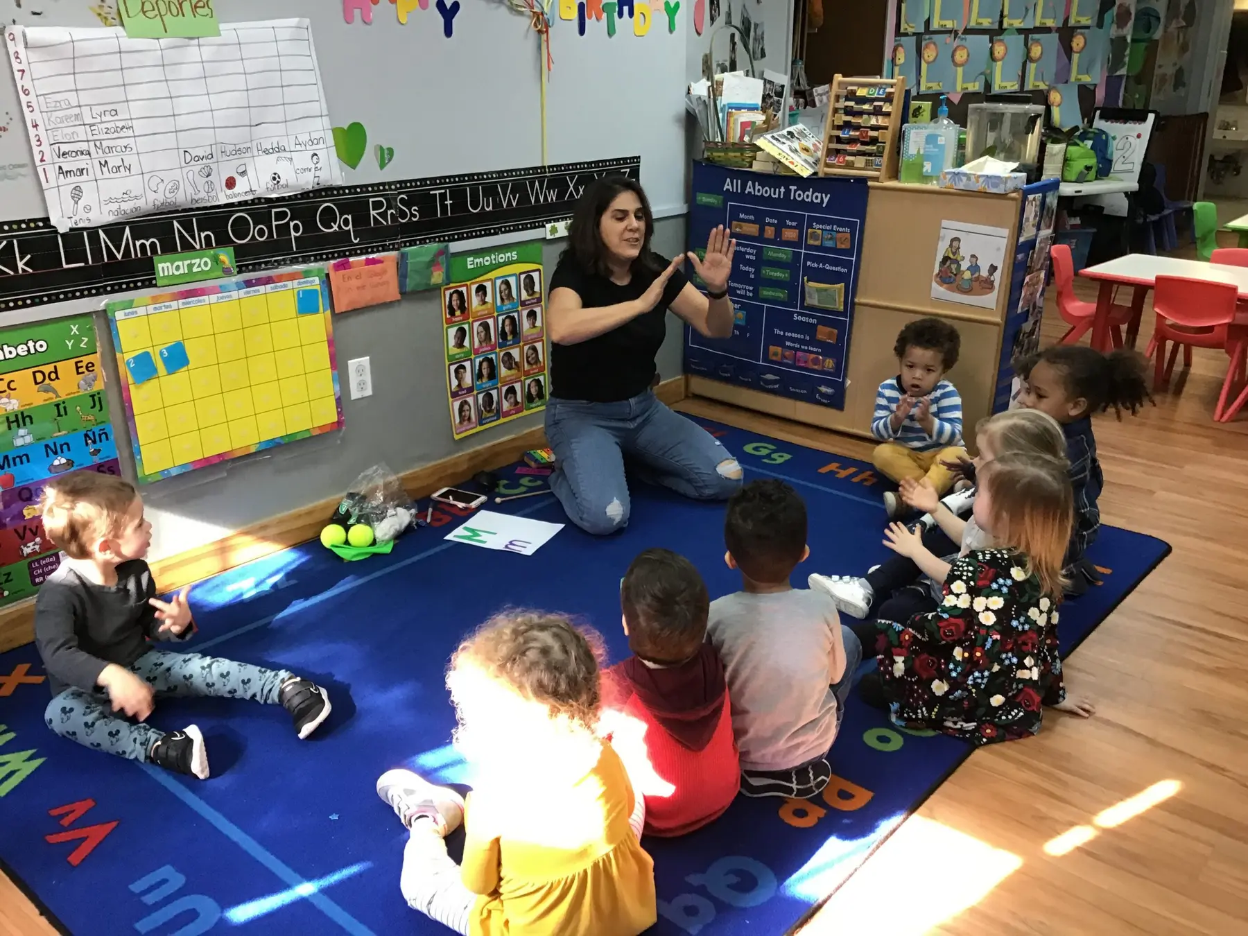 A woman kneels on a blue alphabet rug gesturing with her hands while eight children sit around her in a classroom with wall displays and shelves.