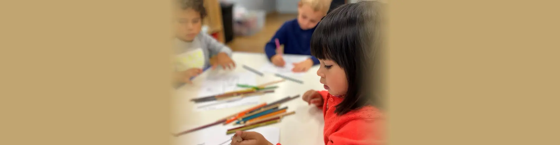 Three young children sit at a white table drawing with colored pencils on paper.