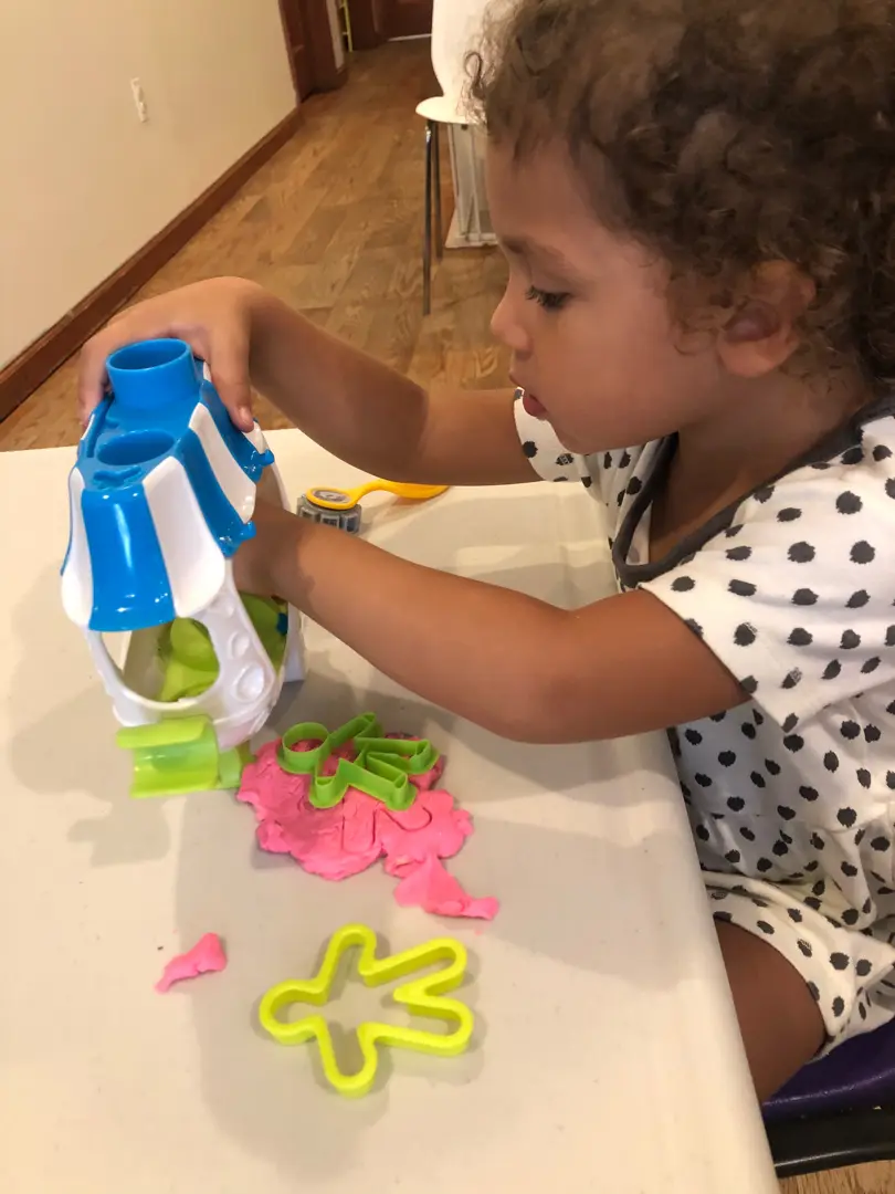 A child with curly hair sits at a white table and plays with a blue and white toy structure alongside pink and green modeling dough and a yellow cutter.