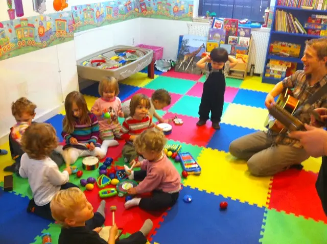 A group of toddlers sits on colorful floor mats playing with musical instruments while an adult plays guitar nearby.