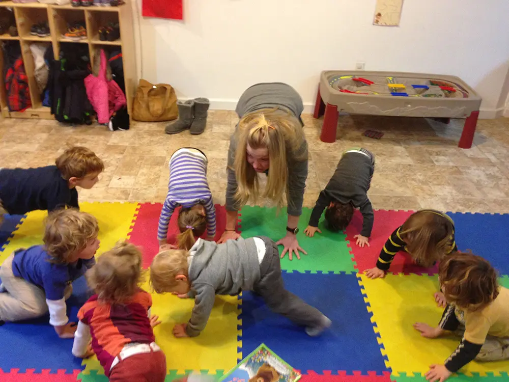 A group of toddlers and an adult on colorful floor mats doing a crawling exercise in a childcare room with toys and storage units.