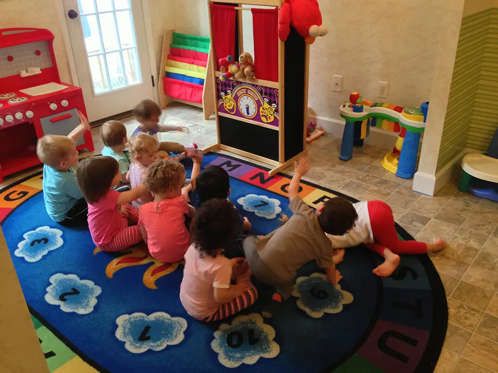 Nine preschoolers sit on a colorful rug with numbers and letters, interacting around a play kitchen and toy theater in a bright classroom.