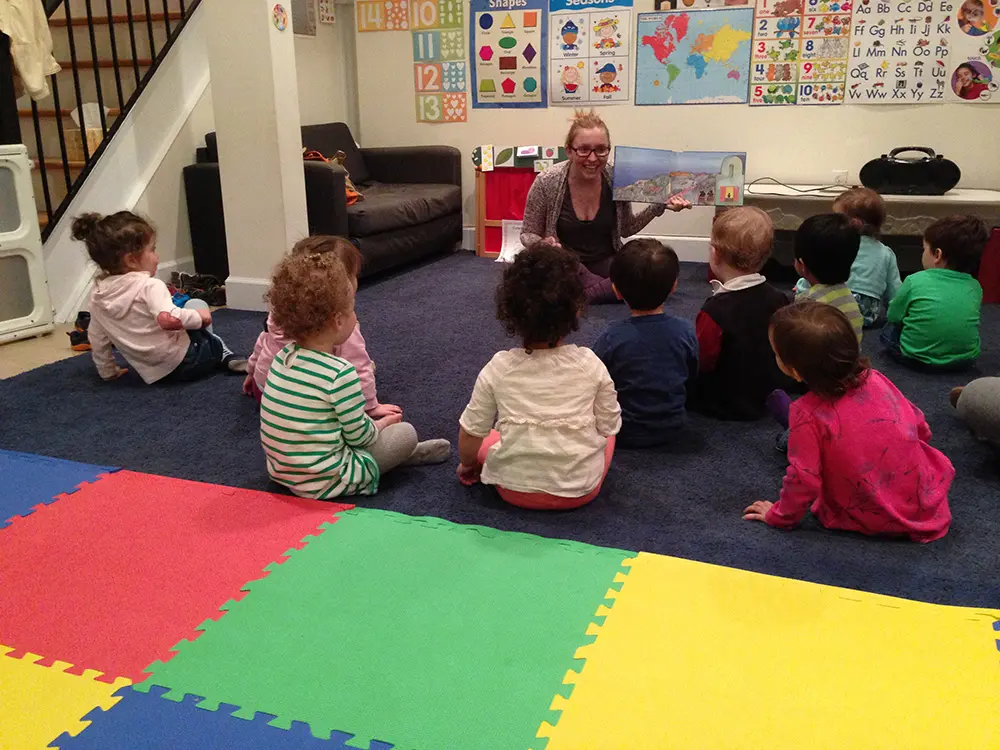 A group of preschool children sitting on a colorful rug in a classroom while an adult holds up a book.