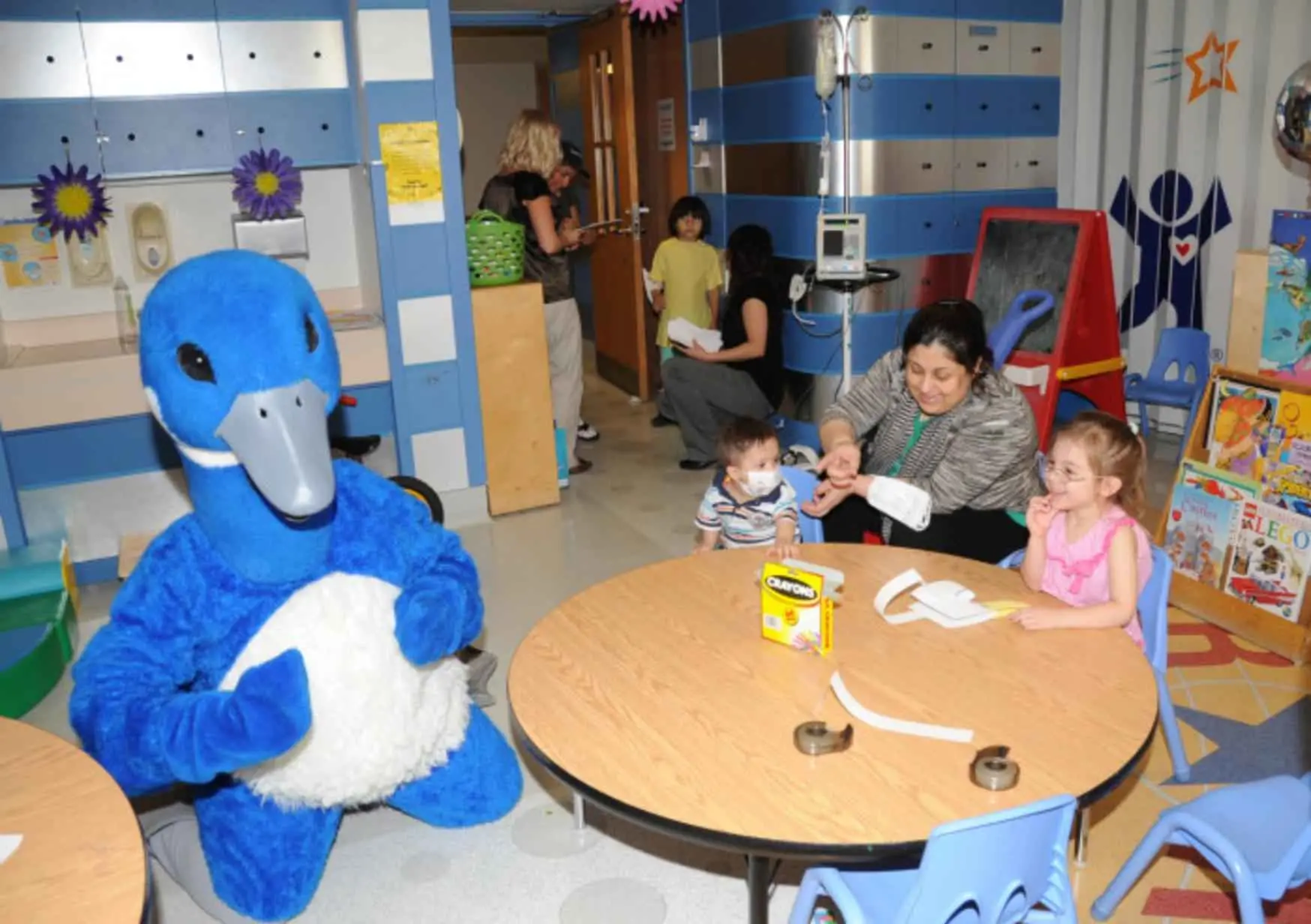 A blue mascot stands near a table with children and adults in a colorful indoor childcare facility.