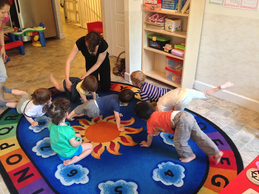 Seven toddlers and one adult gather around a colorful sun-shaped rug in a classroom setting.
