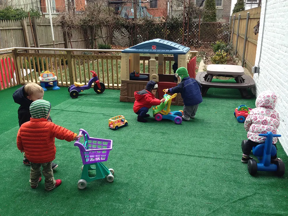 Six toddlers play with toy shopping cart, tricycle, and ride-on toys on green artificial turf in a backyard playground.