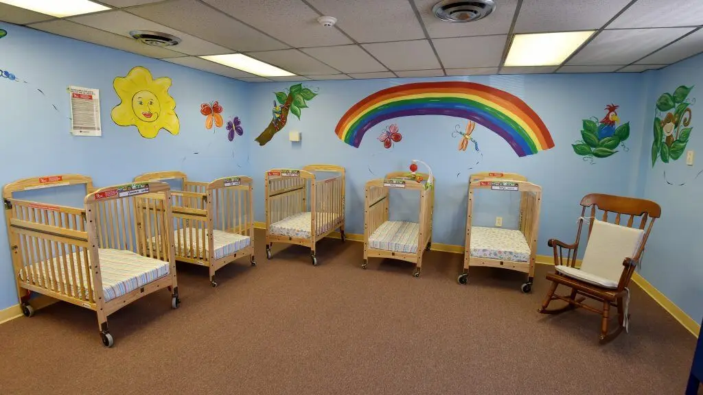 A nursery room with multiple wooden cribs on brown carpet and colorful wall murals including a sun, rainbow, butterflies, and animals.