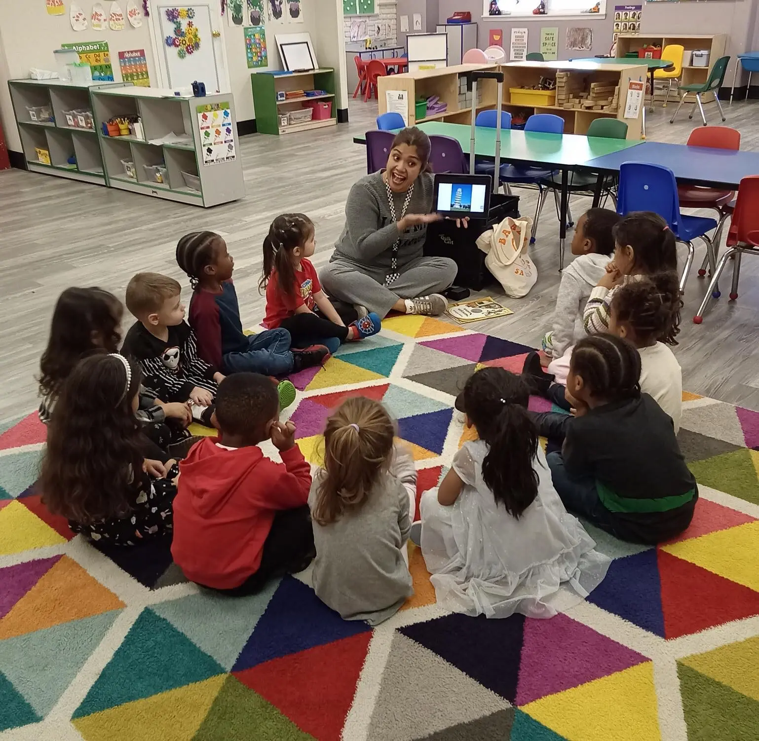 A teacher sits on a colorful rug with a group of preschoolers, holding a tablet while children sit in a circle around her.