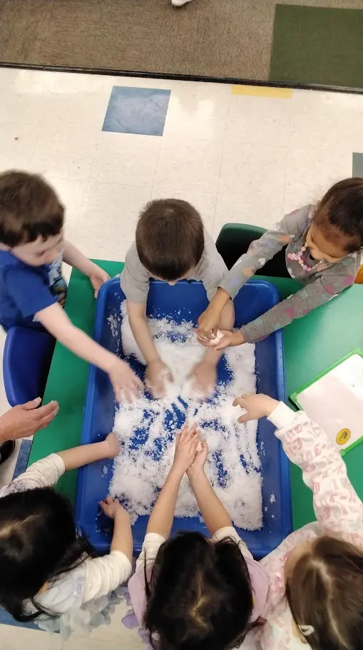 Five children and one adult hand interact with white material in a blue tray on a green table.