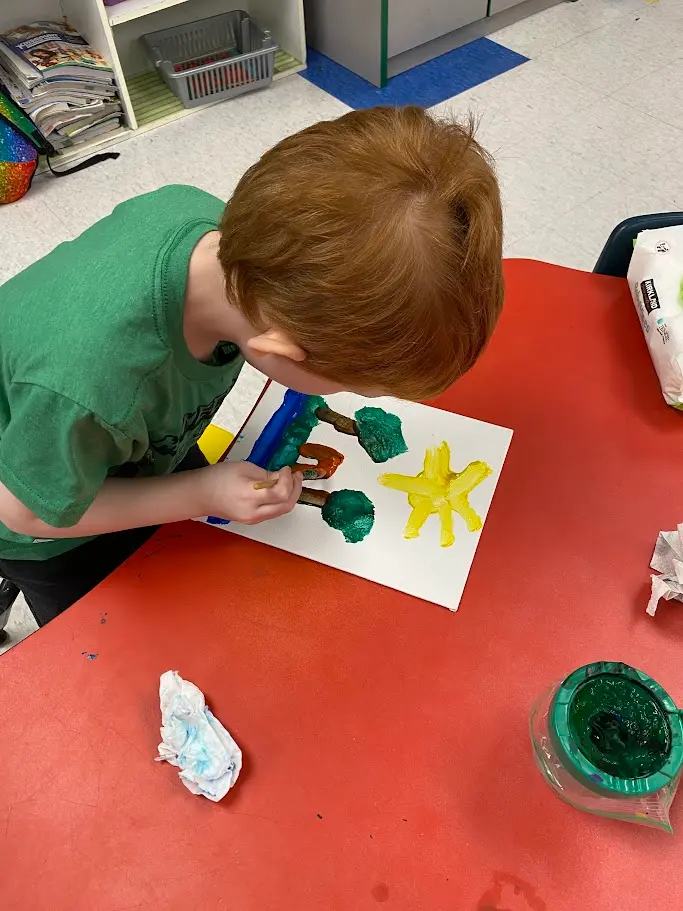 A child in a green shirt paints on a white paper with a yellow sun and green trees on a red table.