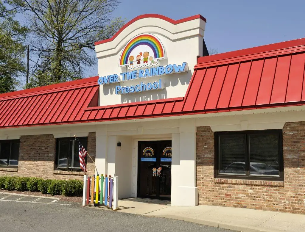A preschool building with a red roof, rainbow sign, American flag, and colorful poles outside.