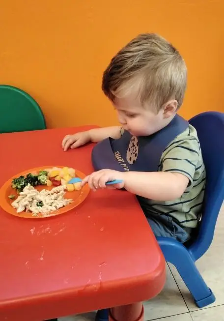 A toddler sits at a red table with an orange plate of food using blue utensils.