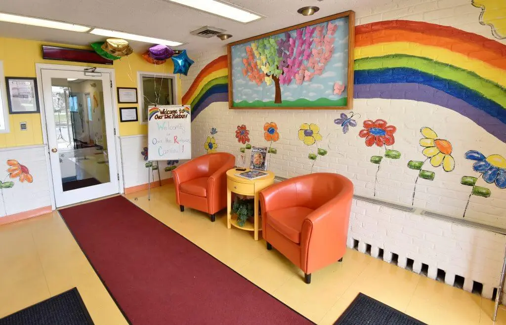 A colorful childcare facility lobby with orange chairs, a rainbow mural, flowers on the wall, and a welcome sign.