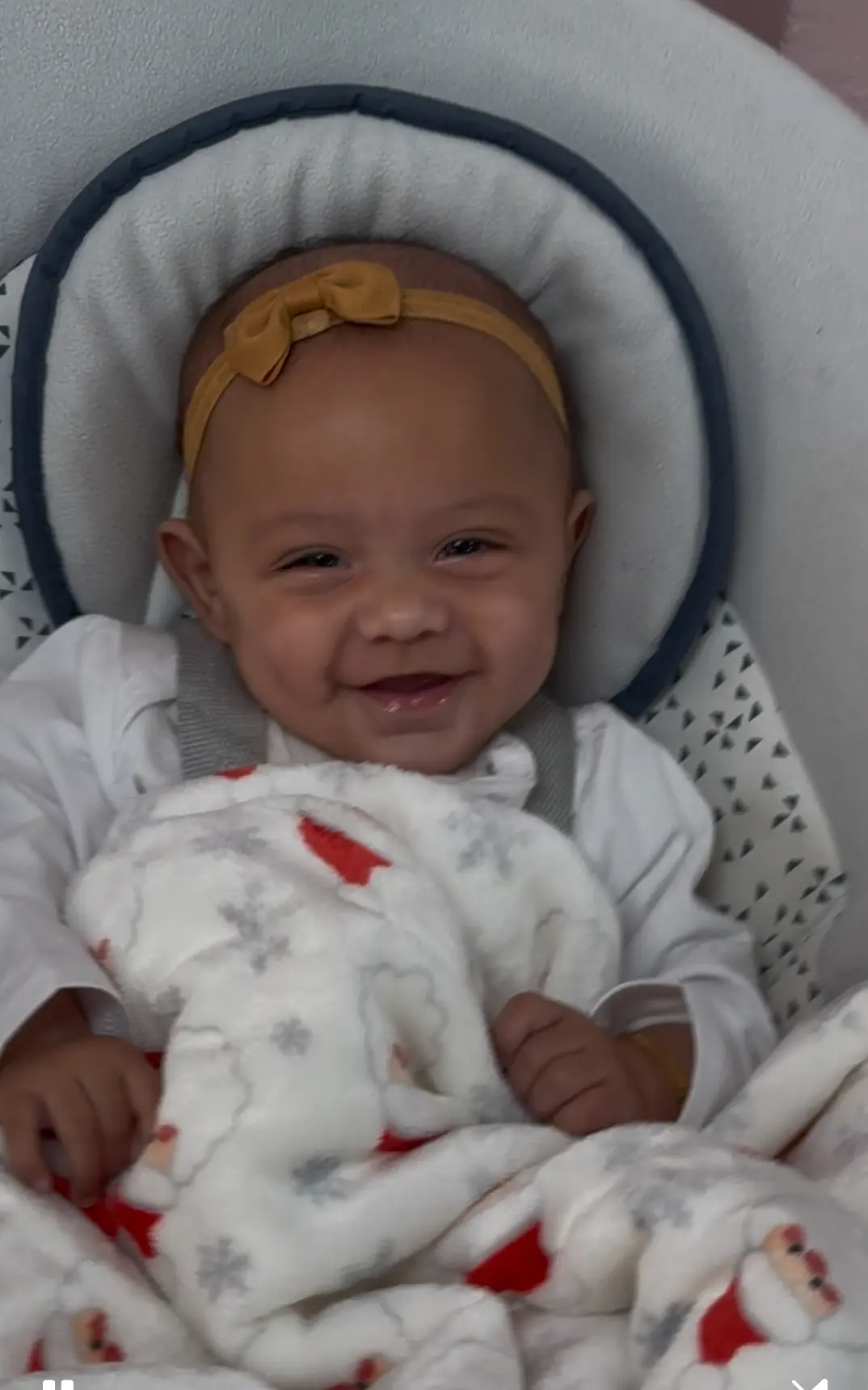 A smiling baby wearing a yellow bow headband sits in a bouncer seat wrapped in a white blanket with red and gray patterns.