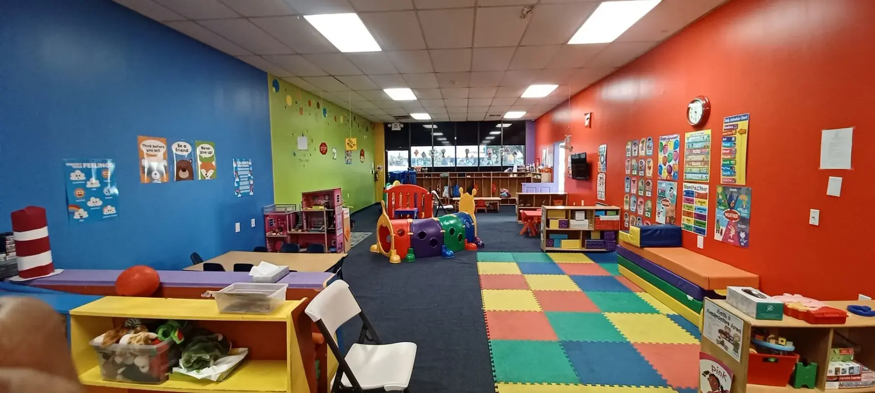 An empty childcare classroom with blue, green, and red walls, colorful floor mats, toy shelves, and play equipment.