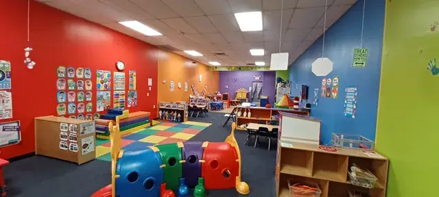 A brightly colored childcare room with red, blue, and green walls, containing toy storage units, a patterned rug, small tables, chairs, and play structures.