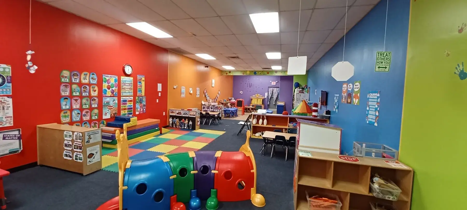 A brightly colored childcare room with red, orange, blue, and green walls, containing a large plastic tunnel toy, foam mats, wooden storage shelves, and small tables with chairs.