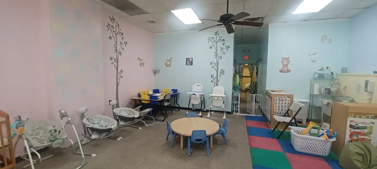 An empty childcare room with pastel walls, tree decals, a small table with blue chairs, several infant bouncers and high chairs, and a colorful foam play mat.