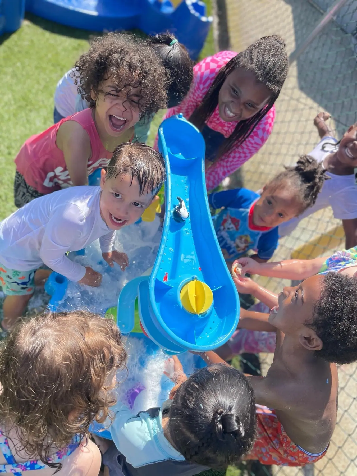 A group of children gather around a blue water play table, with one child smiling at the camera while others reach into the splashing water.