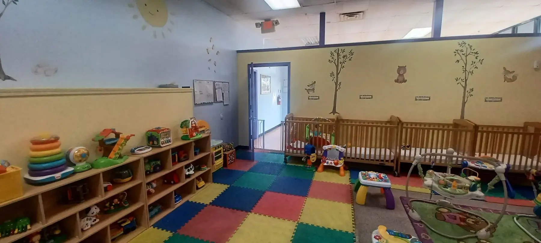 A childcare room with wooden cubby shelves holding toys, colorful foam floor mats, and a row of wooden cribs along a wall decorated with tree and animal decals.