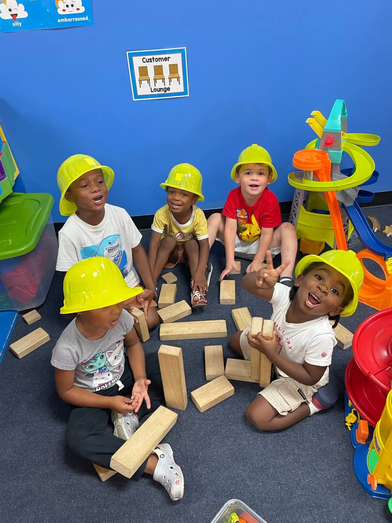 Five children wearing yellow hard hats sit on a blue carpet playing with wooden blocks and a toy marble run in a classroom.