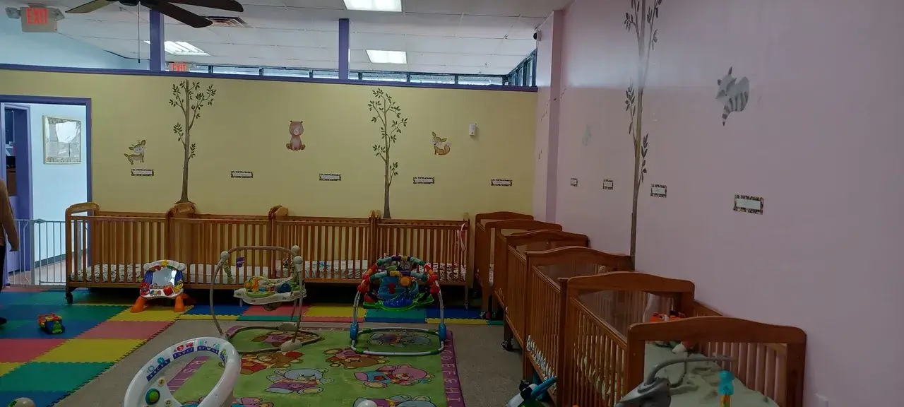 A childcare room with wooden cribs lining the walls, colorful floor mats, and baby activity centers on the floor.