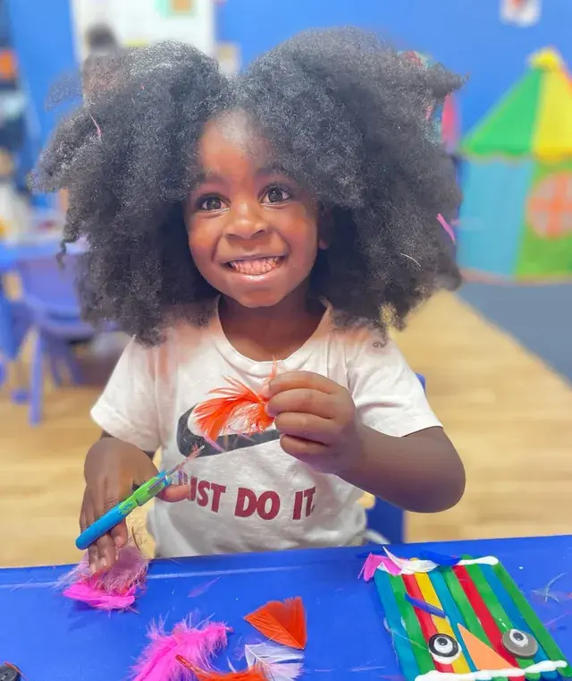 A smiling child with voluminous hair holds an orange feather while sitting at a blue table with craft supplies including feathers, a painted stick craft, and a paintbrush.