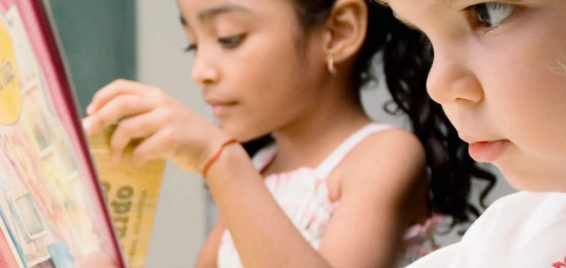 Two girls look at a book with a red cover, one holding a page while the other watches closely.