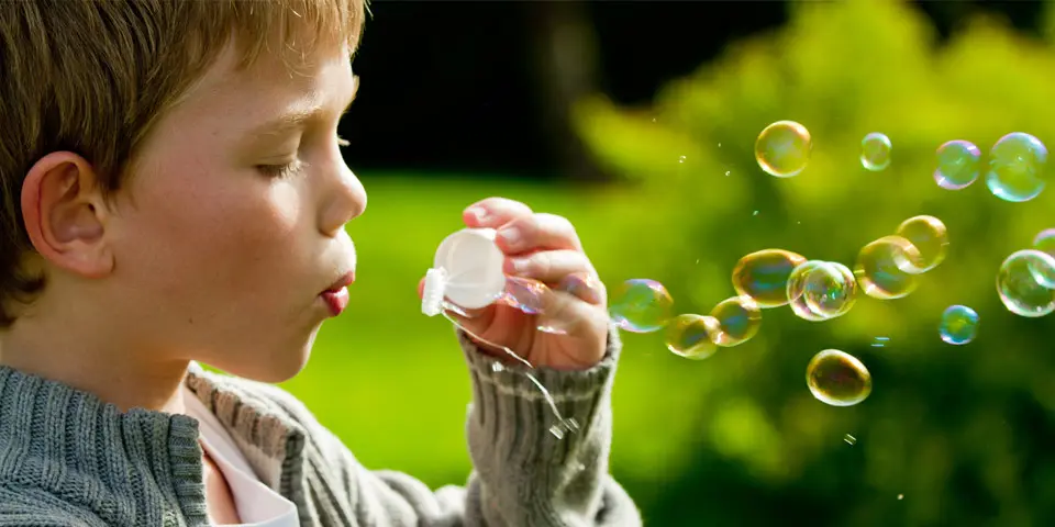 A young boy blows bubbles through a white plastic wand while standing outside against a blurred green background.
