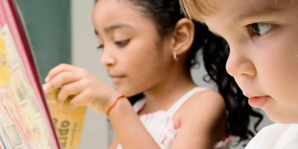 Two young girls look closely at a colorful book, with one girl touching a page while the other watches.