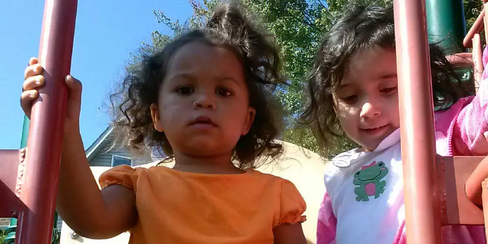 Two young girls stand on a playground structure, one in an orange shirt holding a red pole and another in a white shirt with a frog graphic beside her.
