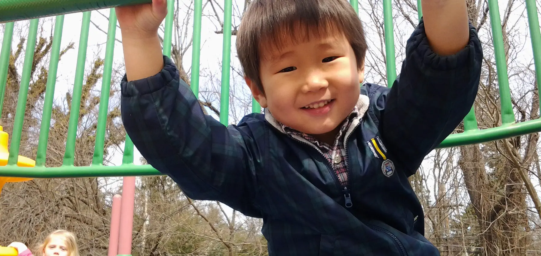 A young boy in a dark plaid jacket hangs from a green metal play structure while smiling, with another child visible in the lower left background.