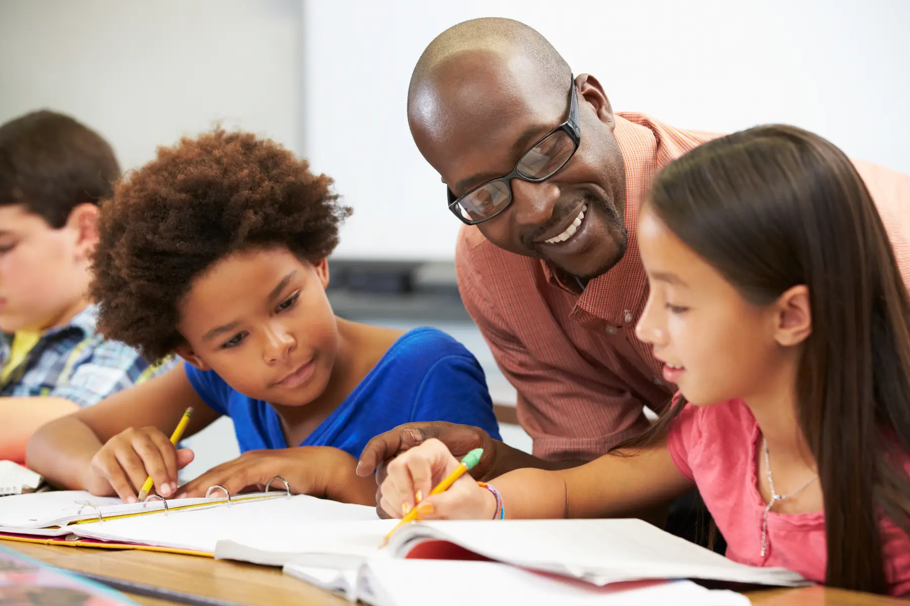A man in glasses leans over two children writing in notebooks with pencils at a table while another child sits at left.