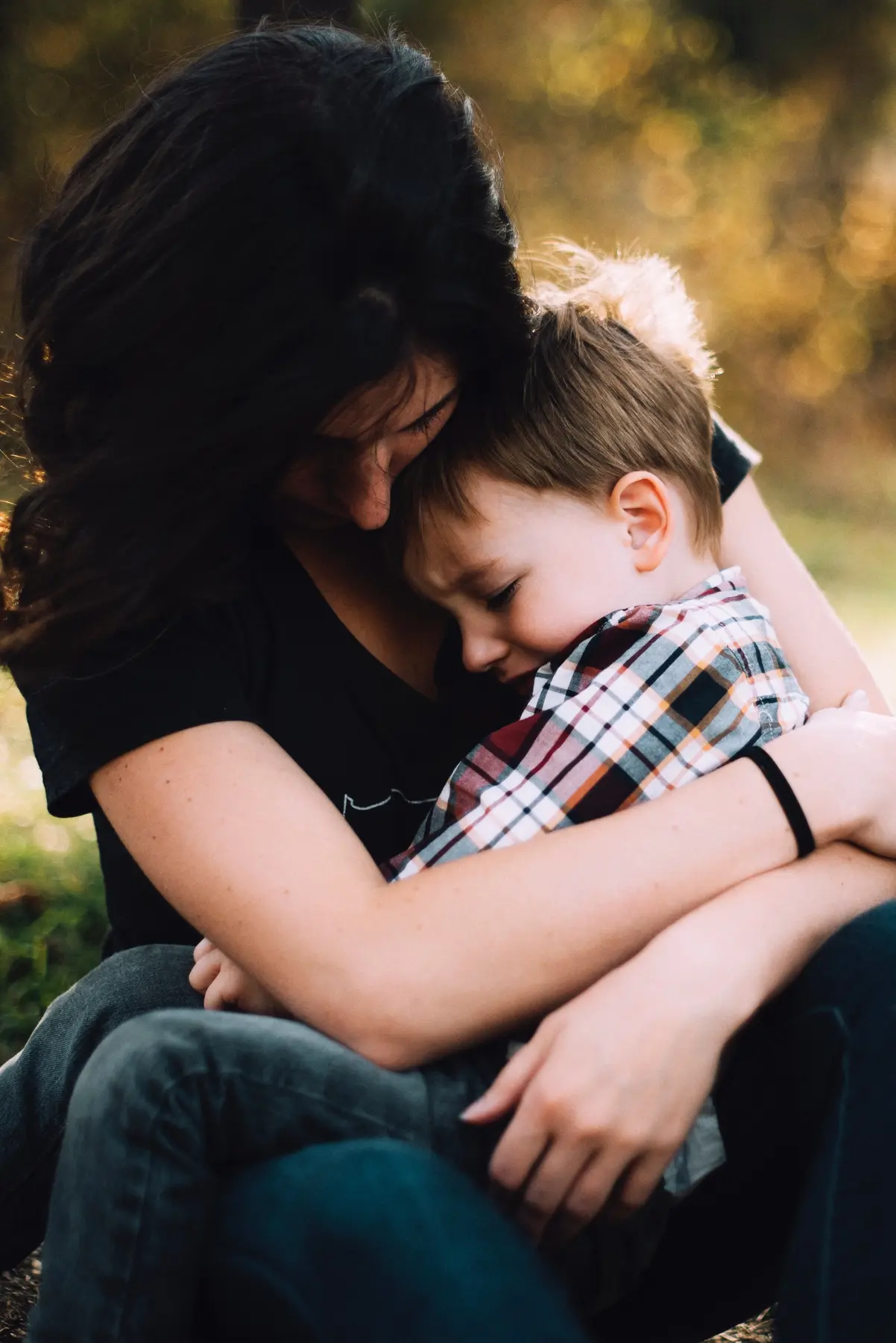 A person with long dark hair embraces a young child wearing a plaid shirt, both seated outdoors with blurred foliage behind them.