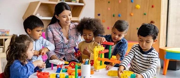 A woman sits at a table with five children who are playing with colorful building blocks and toys.