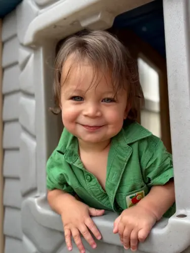 A young child in a green shirt leans on the ledge of a gray plastic play structure and smiles at the camera.