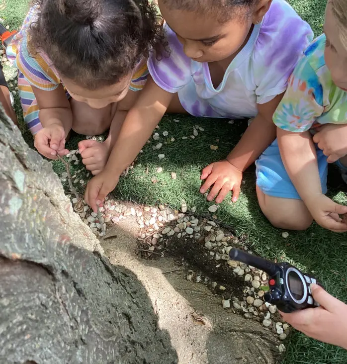 Three children crouch on artificial grass near a tree trunk, examining small objects on the ground while an adult hand holds a black two-way radio nearby.