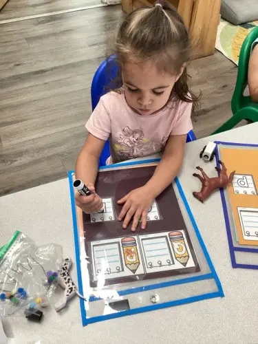 A young girl sits at a table and uses a marker on a laminated worksheet while touching the paper with her other hand.