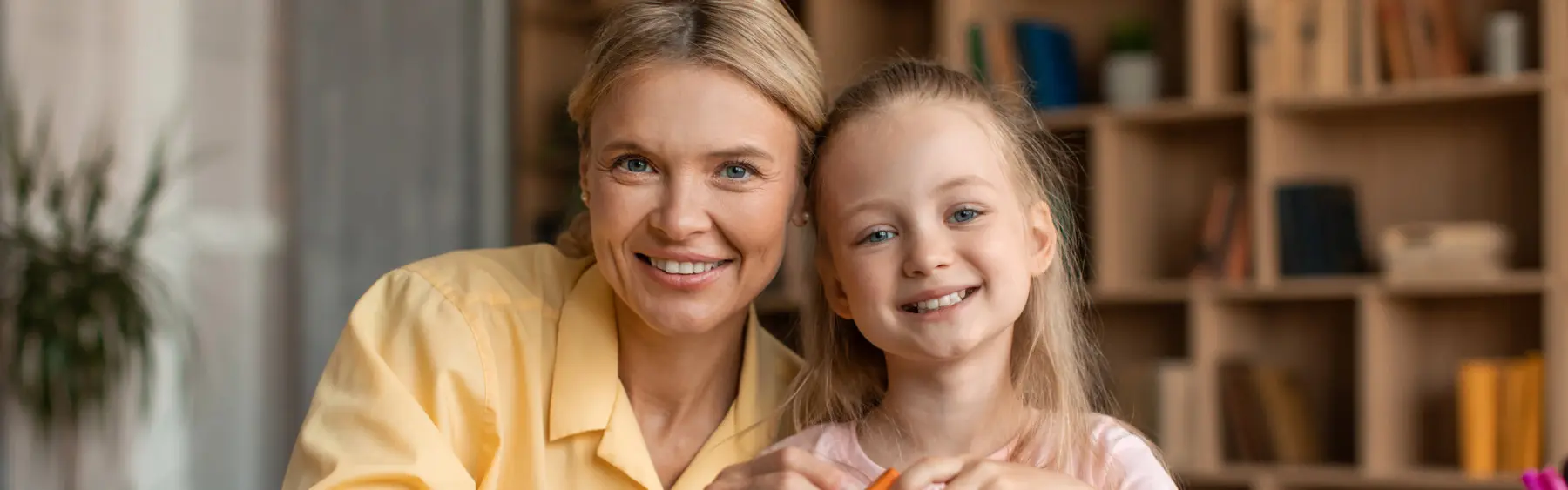 A woman in a yellow shirt and a girl in a pink top smile at the camera while holding a pencil over a surface in front of a wooden shelf.