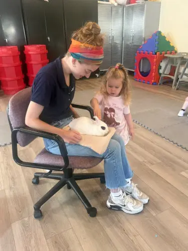 A woman seated in a rolling chair holds a white rabbit on a paper bag while a young girl stands beside her and reaches toward the animal.