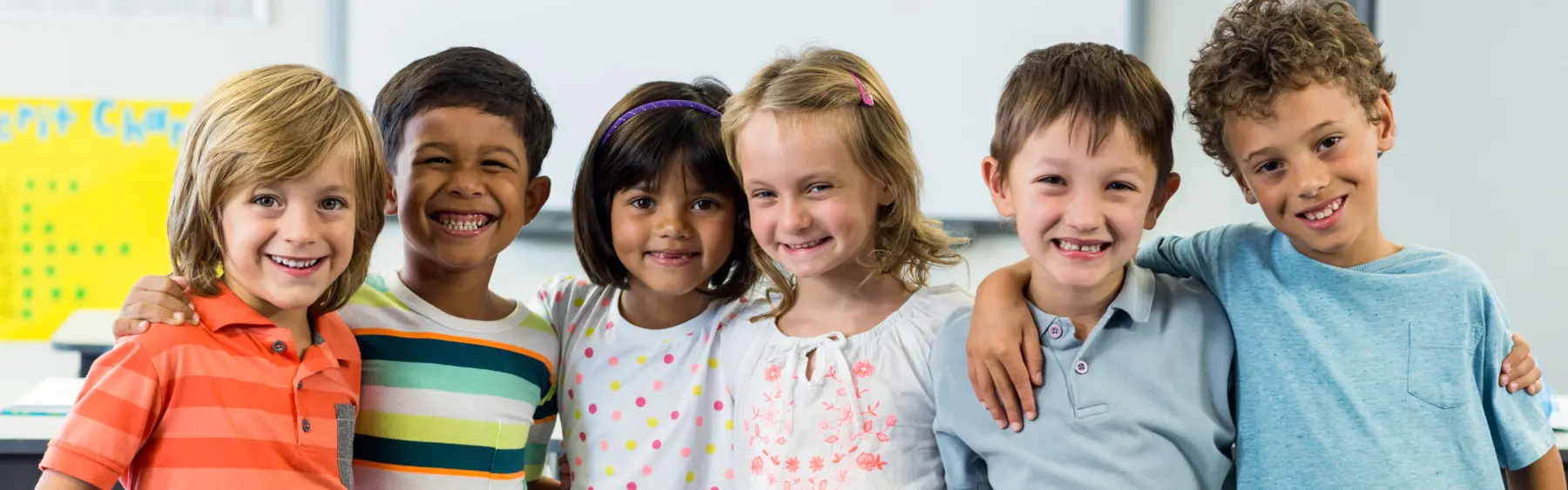 Six children stand shoulder to shoulder smiling at the camera in front of a whiteboard with a yellow chart.