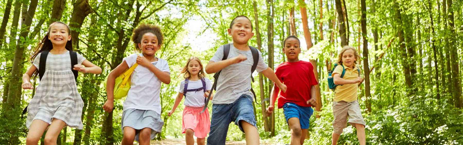 Six children wearing backpacks run toward the camera on a path through a green forest.
