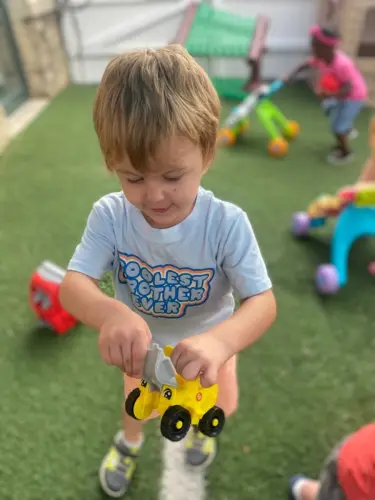 A young boy in a blue shirt stands on green turf holding a yellow toy truck while another child plays with a ride-on toy in the background.