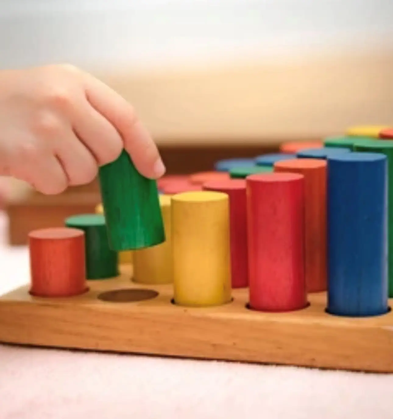 A hand places a green cylindrical peg into a wooden base while other red, yellow, blue, and green pegs stand in the background.